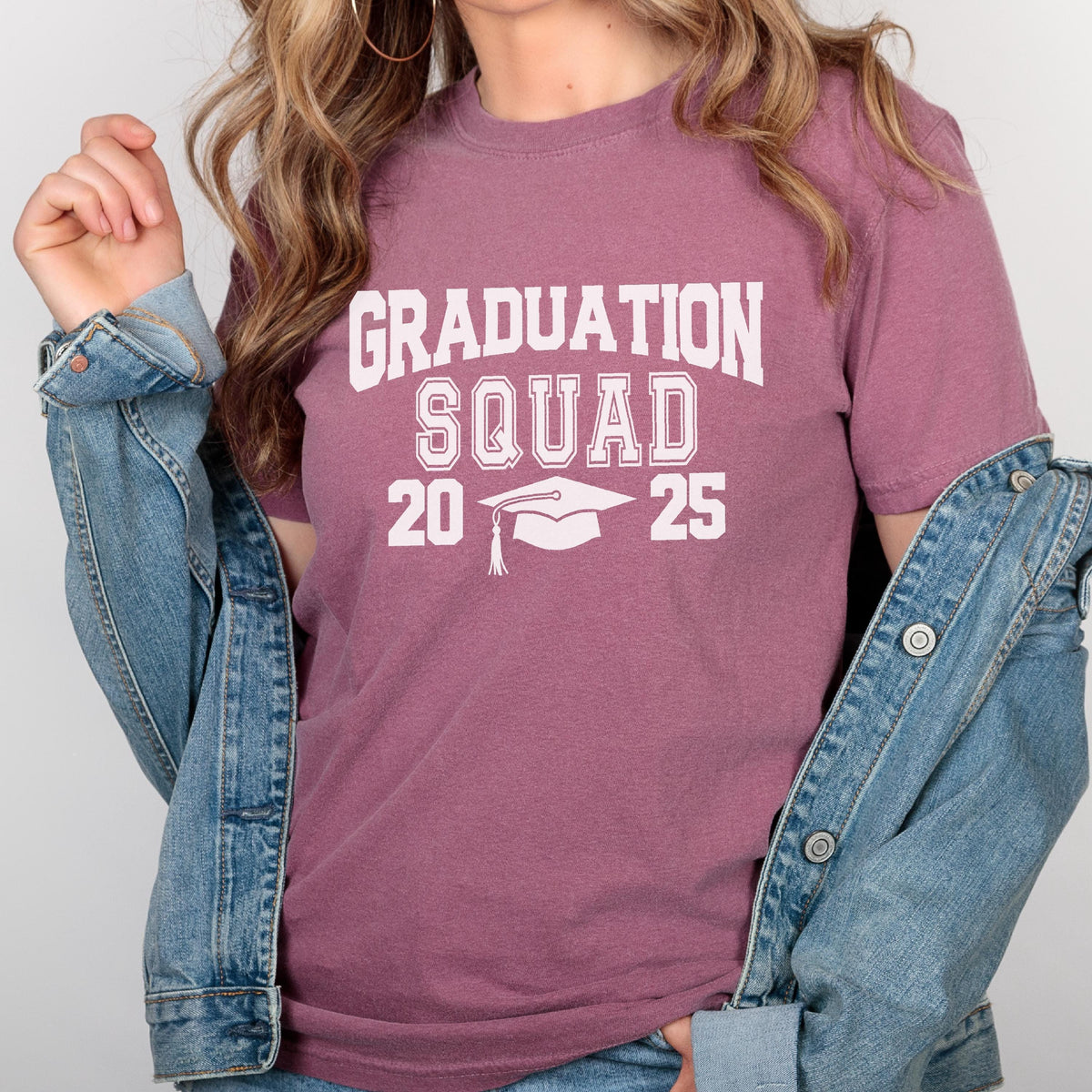 May include: A mauve-colored t-shirt with the words "GRADUATION SQUAD 20 25" printed in white. The design includes a graduation cap. The shirt is paired with a denim jacket.