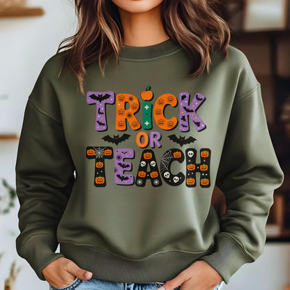 May include: An olive green sweatshirt with the words "TRICK OR TEACH" in a colorful Halloween-themed design. The letters are decorated with bats, pumpkins, and spiderwebs. The sweatshirt is a crew neck style.