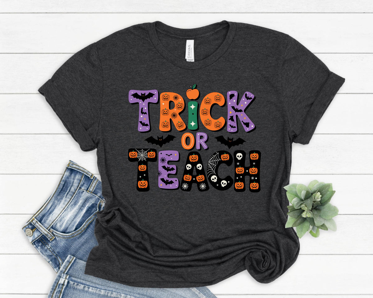 May include: Dark gray t-shirt with the words "Trick or Teach" in a colorful Halloween-themed design. The text includes pumpkins, bats, and spiderwebs. The shirt is folded with a pair of blue jeans.