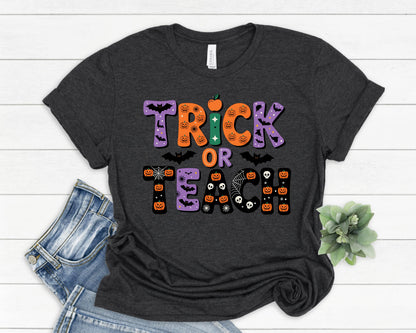 May include: Dark gray t-shirt with the words "Trick or Teach" in a colorful Halloween-themed design. The text includes pumpkins, bats, and spiderwebs. The shirt is folded with a pair of blue jeans.
