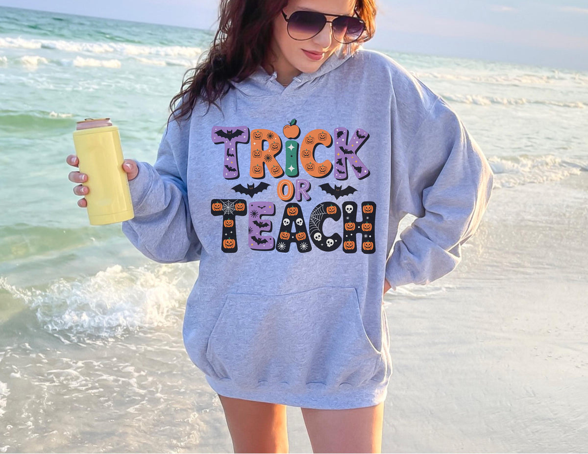 May include: A light gray hoodie with the words "Trick or Teach" in a colorful Halloween-themed design. The design includes pumpkins, bats, and spiderwebs. The hoodie has a front pocket and a drawstring hood.