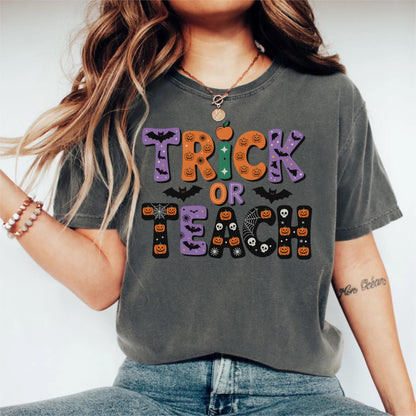 May include: Gray t-shirt with the words "Trick or Teach" in a Halloween-themed design. The text features orange pumpkins, purple bats, and a green apple. The shirt is a comfortable, casual style.
