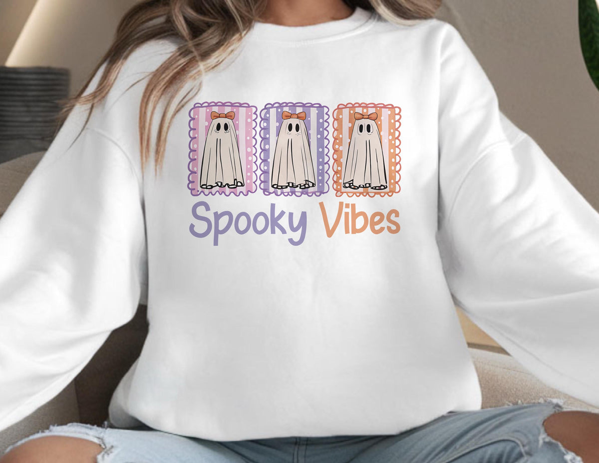 May include: White sweatshirt featuring three cartoon ghosts in decorative frames. Each ghost has a bow and is set against a striped background. The phrase "Spooky Vibes" is printed in purple and orange below the ghosts.