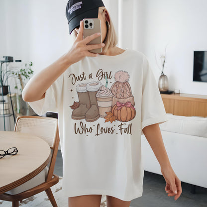 May include: Cream-colored t-shirt with a fall-themed graphic. The graphic includes boots, a coffee cup, a knit hat, and a pumpkin. The text on the shirt reads "Just a Girl Who Loves Fall."