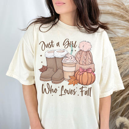 May include: Cream-colored t-shirt with a fall-themed graphic. The graphic includes brown boots, a coffee cup, a knit hat, and a pumpkin. The text "Just a Girl Who Loves Fall" is printed below the graphic.