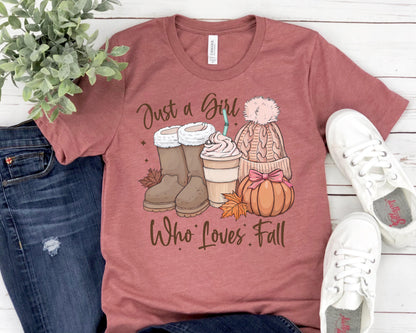May include: A maroon t-shirt with the text "Just a Girl Who Loves Fall." The graphic includes brown boots, a coffee drink, a knit hat with a pink pom-pom, and a pumpkin. White sneakers are on the right.