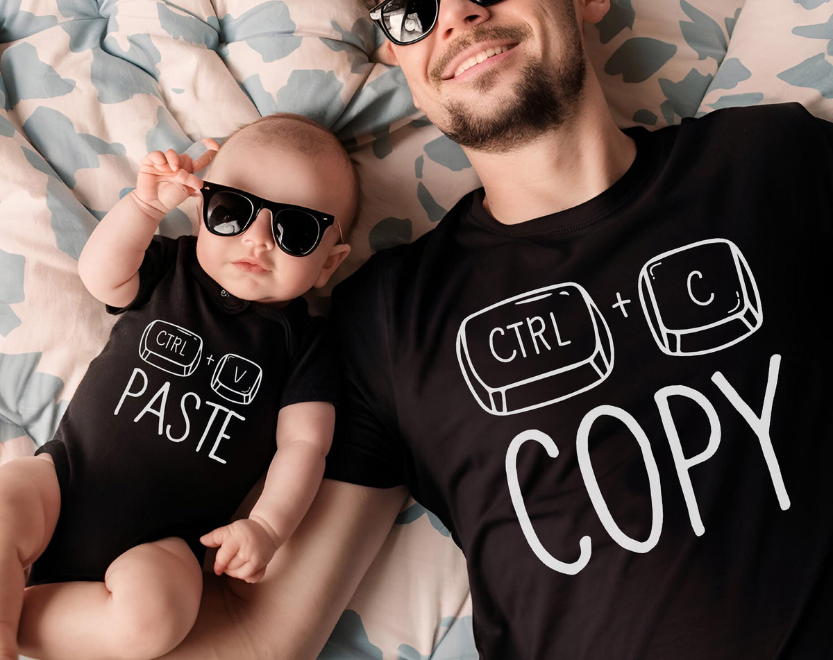 May include: A black onesie with the text "CTRL + PASTE" and a black t-shirt with "CTRL + COPY" printed in white. Both the baby and the adult are wearing black sunglasses. The image is taken from above.