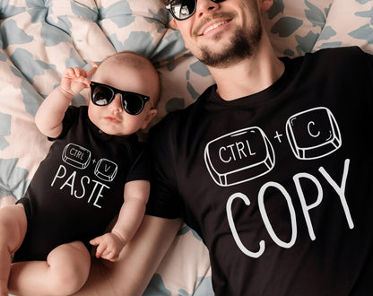 May include: A black onesie with the text "CTRL + PASTE" and a black t-shirt with "CTRL + COPY" printed in white. Both the baby and the adult are wearing black sunglasses. The image is taken from above.