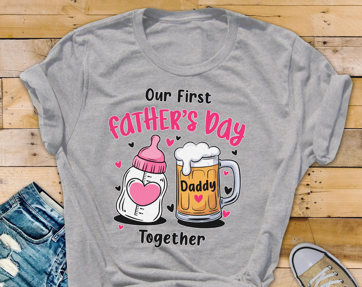 May include: A heather gray t-shirt with a graphic for Father's Day. The design includes a baby bottle with a pink heart and a beer mug with the word "Daddy." The text "Our First Father's Day Together" is also displayed.