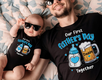 May include: A black t-shirt and baby onesie set with the text "Our First Father's Day" in blue and white. The design includes a baby bottle with a heart and a beer mug with the word "Daddy". Both are wearing sunglasses.