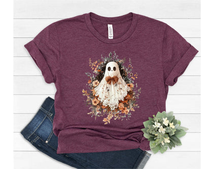 May include: A maroon t-shirt featuring a whimsical ghost design. The ghost is adorned with a brown bow and surrounded by a floral arrangement in shades of orange, brown, and green. The shirt is paired with blue denim jeans.