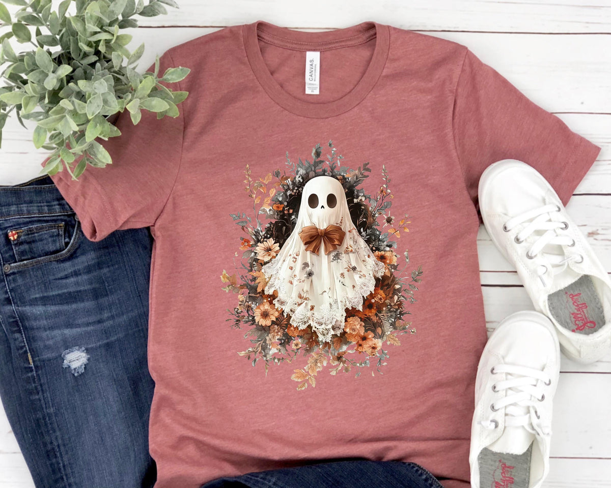 May include: A dusty rose t-shirt featuring a whimsical ghost design. The ghost is adorned with a bow tie and a floral arrangement. The shirt is paired with blue jeans and white sneakers, creating a casual, everyday look.