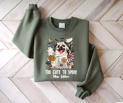 May include: Olive green sweatshirt featuring a graphic of a pug dressed as a mummy, surrounded by flowers, ghosts, and bats. The text "TOO CUTE TO SPOOK Max Edition" is printed below the graphic. The sweatshirt has long sleeves and a crew neck.