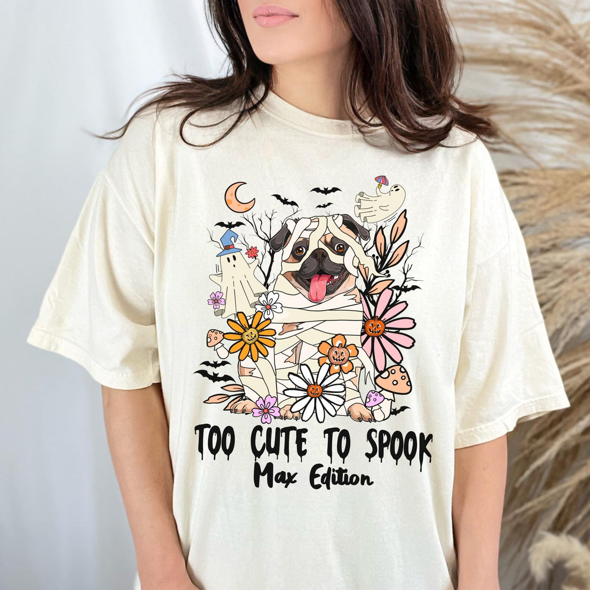 May include: Cream-colored t-shirt featuring a Halloween-themed graphic. The design includes a pug wrapped in bandages, surrounded by flowers, ghosts, bats, and the text "TOO CUTE TO SPOOK Max Edition."