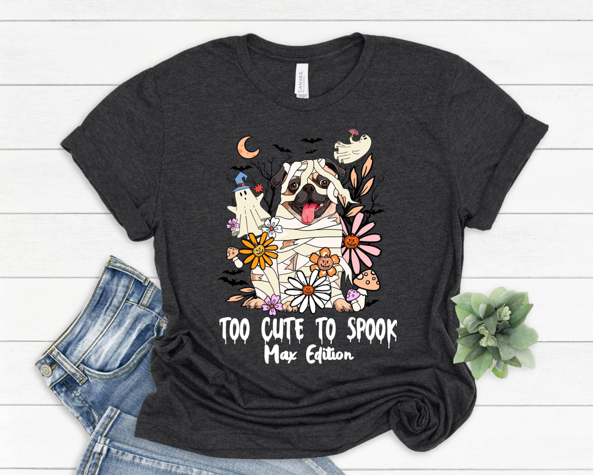 May include: Dark gray t-shirt featuring a pug dressed as a mummy with the text "Too Cute to Spook Max Edition." The design includes flowers, ghosts, bats, and a crescent moon, perfect for Halloween.