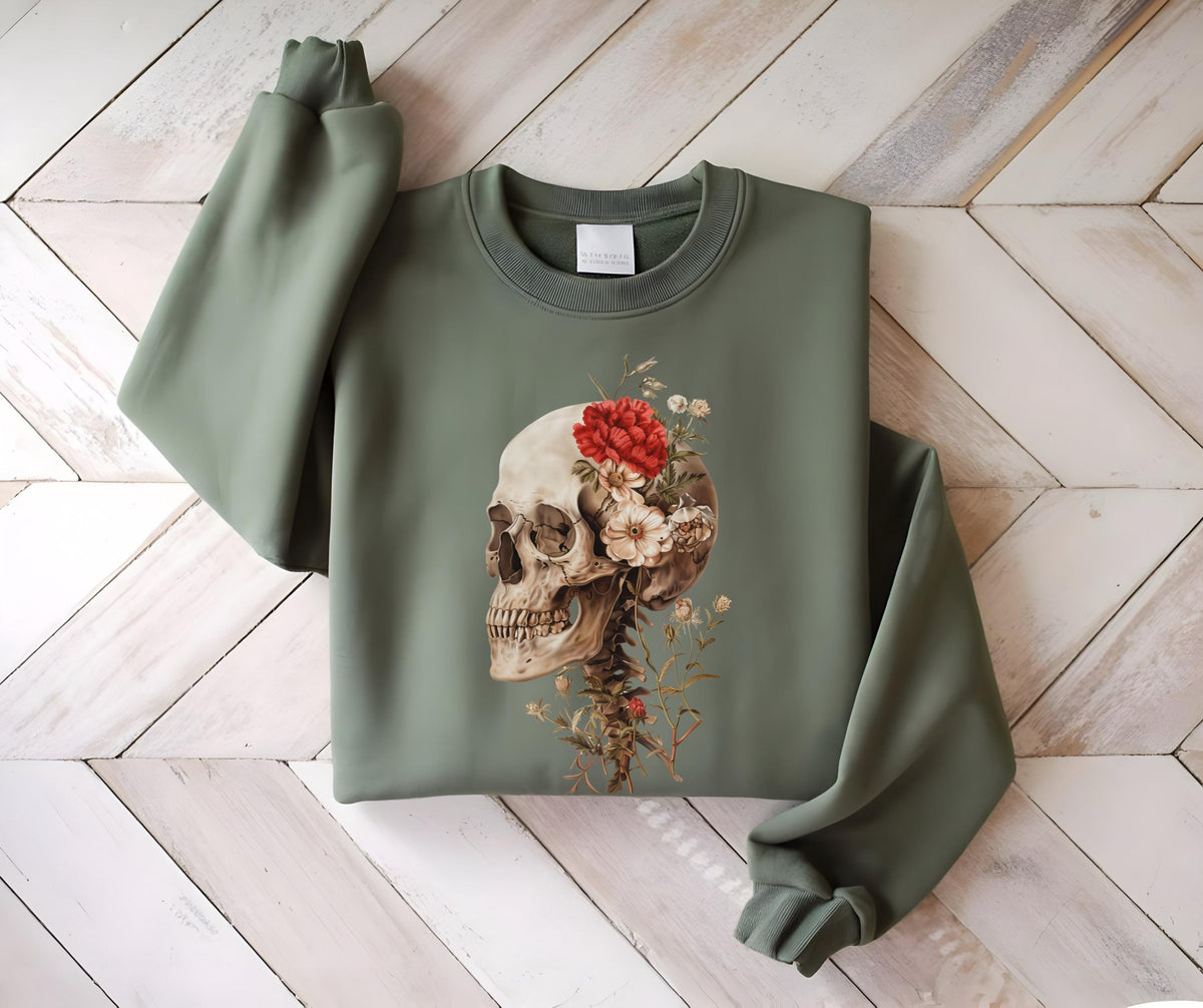 May include: Sage green sweatshirt featuring a detailed illustration of a human skull adorned with red and white flowers. The skull is positioned on the front of the sweatshirt, with the sleeves and body of the garment visible.