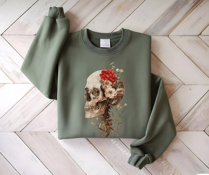 May include: Sage green sweatshirt featuring a detailed illustration of a human skull adorned with red and white flowers. The skull is positioned on the front of the sweatshirt, with the sleeves and body of the garment visible.