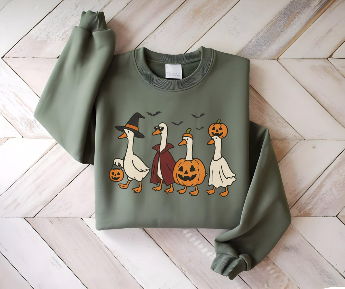 May include: Olive green sweatshirt featuring a Halloween-themed graphic. The design includes four cartoon geese dressed in Halloween costumes: a witch, a vampire, a pumpkin, and a ghost. Bats fly above the geese, and the first goose carries a jack-o'-lantern.