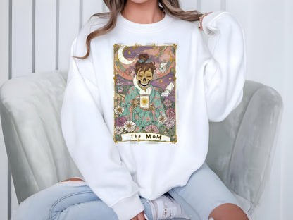 May include: White sweatshirt featuring a tarot card design with a skeleton figure holding a cup with a sun graphic. The card is framed with flowers and the text "The MoM".