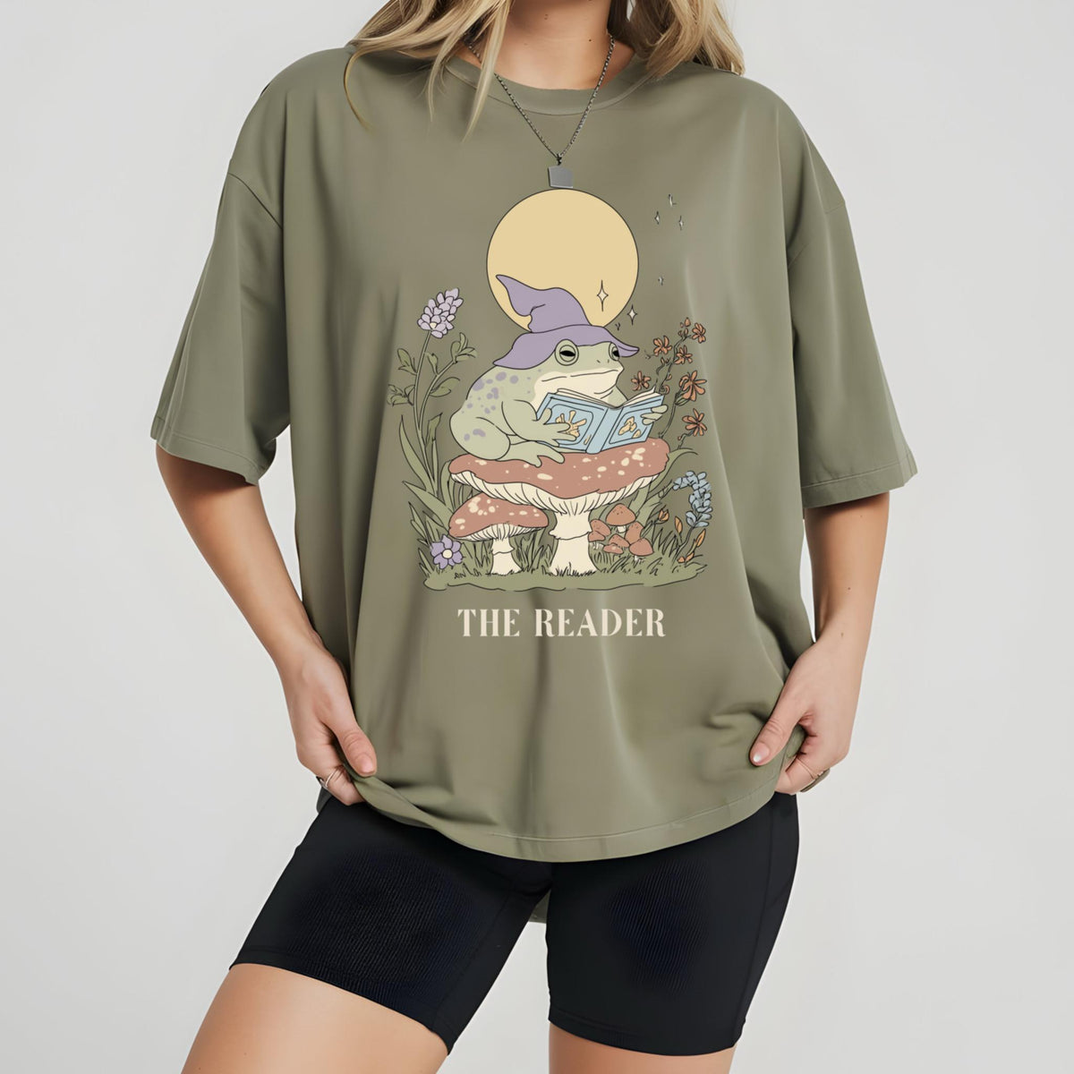 May include: Olive green t-shirt featuring a whimsical illustration of a frog wearing a wizard hat and reading a book while sitting on a mushroom. The text "THE READER" is printed below the illustration. The shirt has short sleeves.