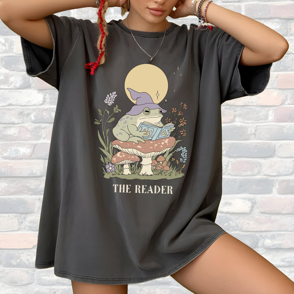 May include: Dark gray t-shirt featuring a whimsical illustration of a frog wearing a wizard hat and reading a book while sitting on a mushroom. The text "THE READER" is printed below the illustration.
