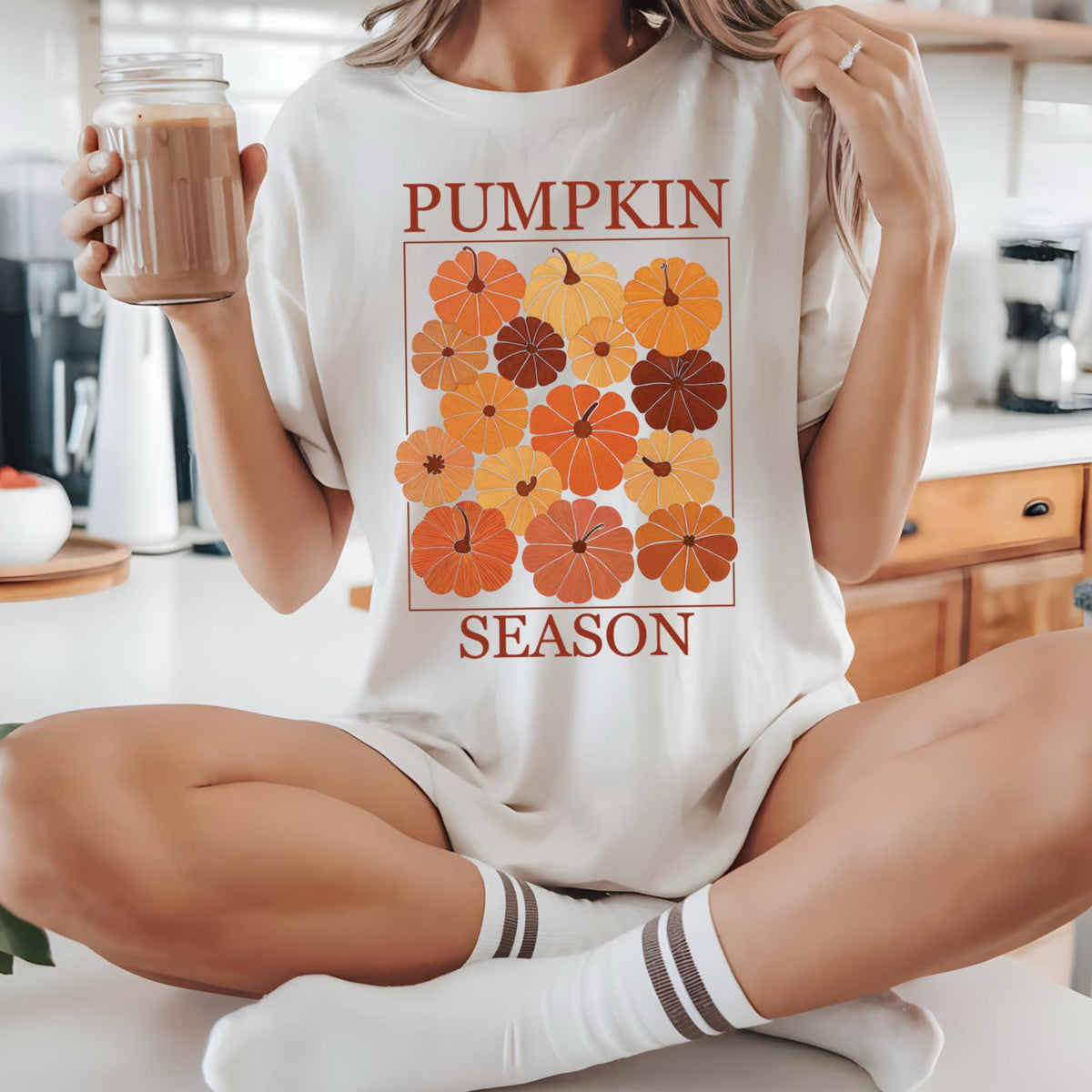 May include: Cream-colored t-shirt with a graphic of pumpkins in shades of orange, yellow, and brown, arranged in a square. The words "PUMPKIN SEASON" are printed above and below the pumpkin graphic in a brown font. The person is wearing white socks with brown stripes.