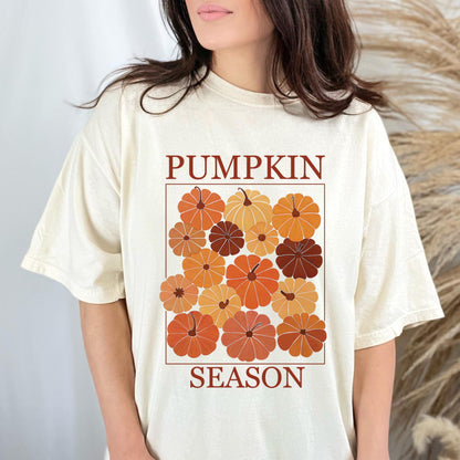May include: Cream-colored t-shirt featuring a rectangular design with the words "PUMPKIN SEASON" in brown. The design includes various orange and brown pumpkins. The shirt has short sleeves and a relaxed fit.