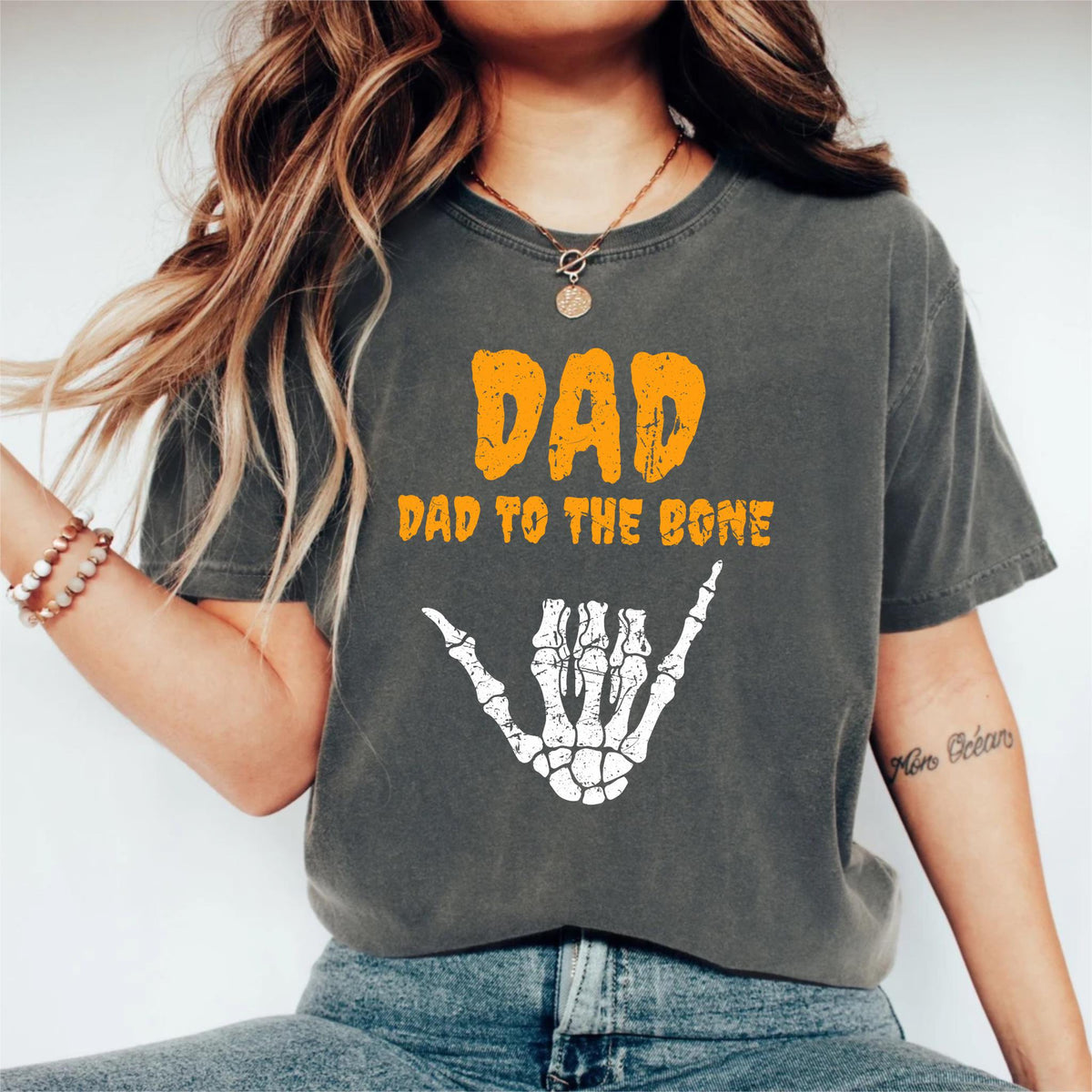 May include: Dark gray t-shirt with the text "DAD" in orange, and "DAD TO THE BONE" below it. A white skeleton hand in the shaka sign is below the text. The shirt is a casual style.