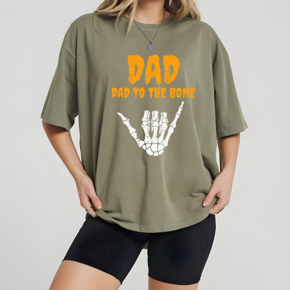 May include: Olive green t-shirt with the text "DAD" in orange, above "DAD TO THE BONE" and a white skeleton hand graphic. The shirt is a casual, oversized fit.