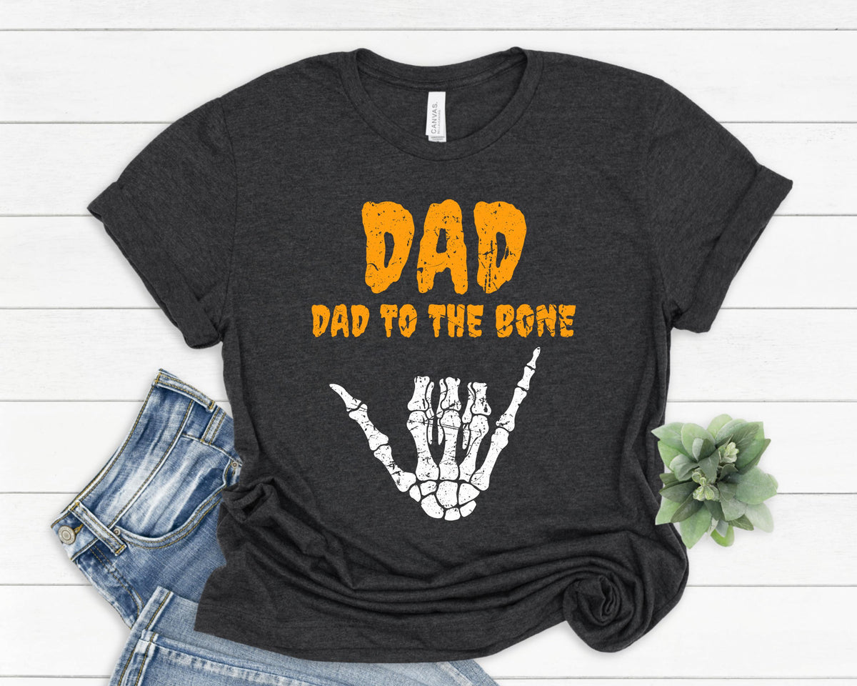 May include: Dark gray t-shirt with the text "DAD" in orange, above "DAD TO THE BONE" in orange. A white skeleton hand in the shaka sign is below the text. The shirt is folded on a white wooden surface.