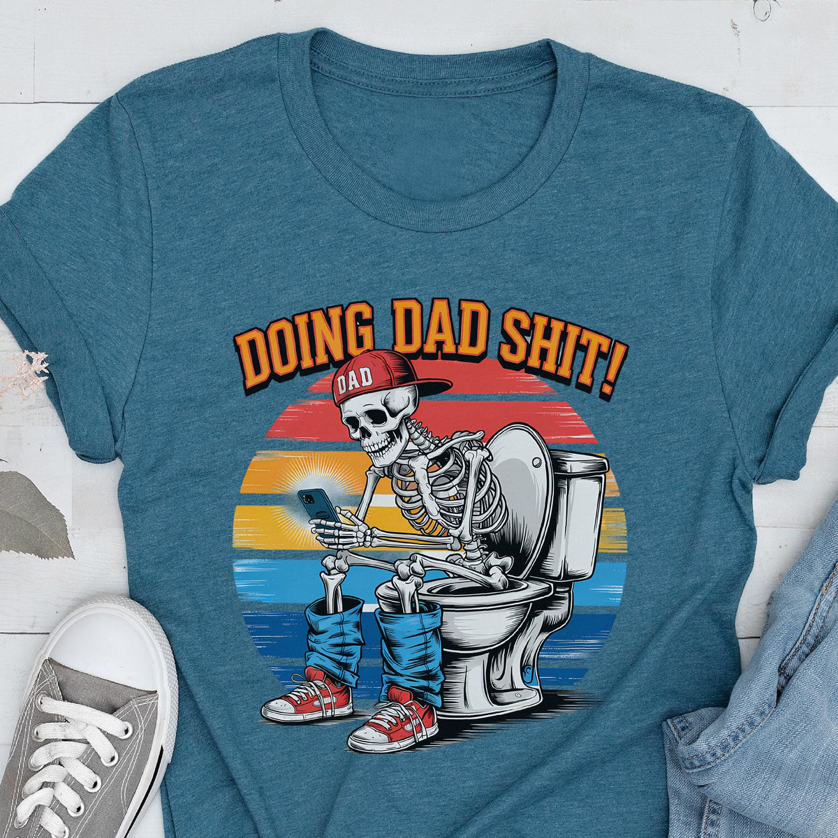 May include: Teal t-shirt with a graphic of a skeleton wearing a red cap and jeans, sitting on a toilet while using a smartphone. The text "DOING DAD SHIT!" is above the skeleton, with "DAD" in a smaller font.