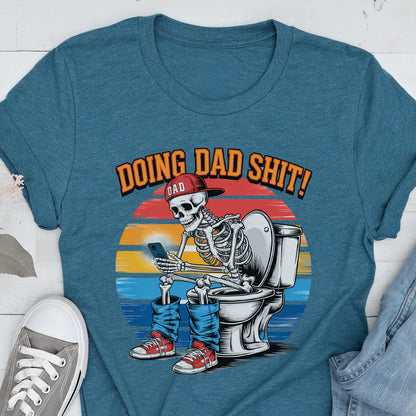 May include: Teal t-shirt with a graphic of a skeleton wearing a red cap and jeans, sitting on a toilet while using a smartphone. The text "DOING DAD SHIT!" is above the skeleton, with "DAD" in a smaller font.