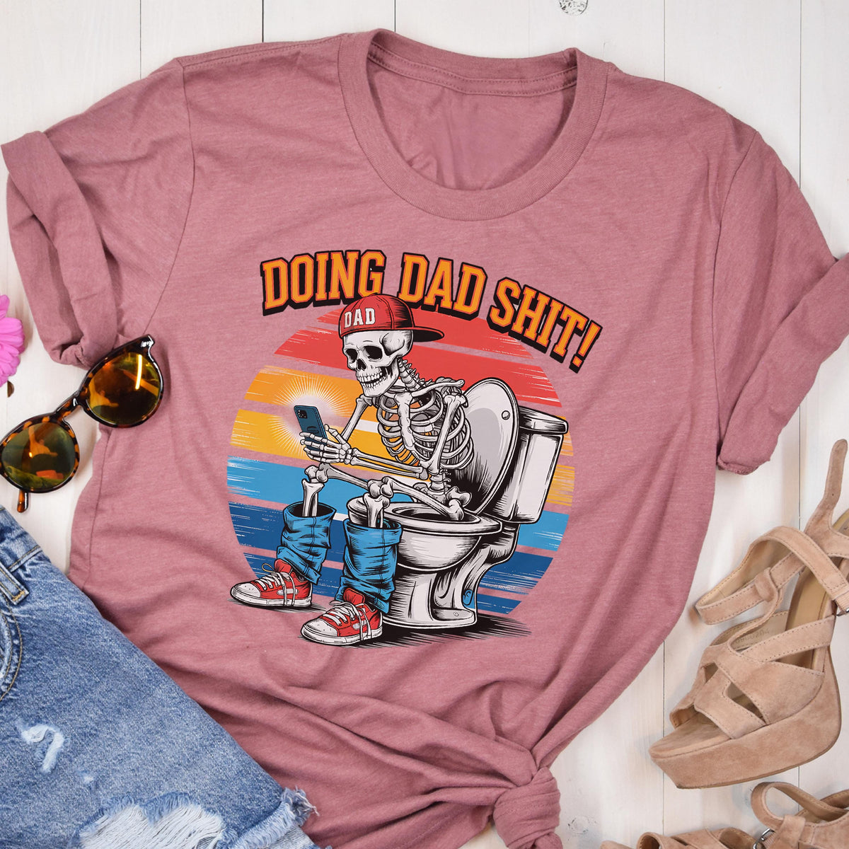May include: A dusty rose t-shirt featuring a graphic of a skeleton wearing a baseball cap and jeans, sitting on a toilet while using a smartphone. The text "DOING DAD SHIT!" is above the graphic.