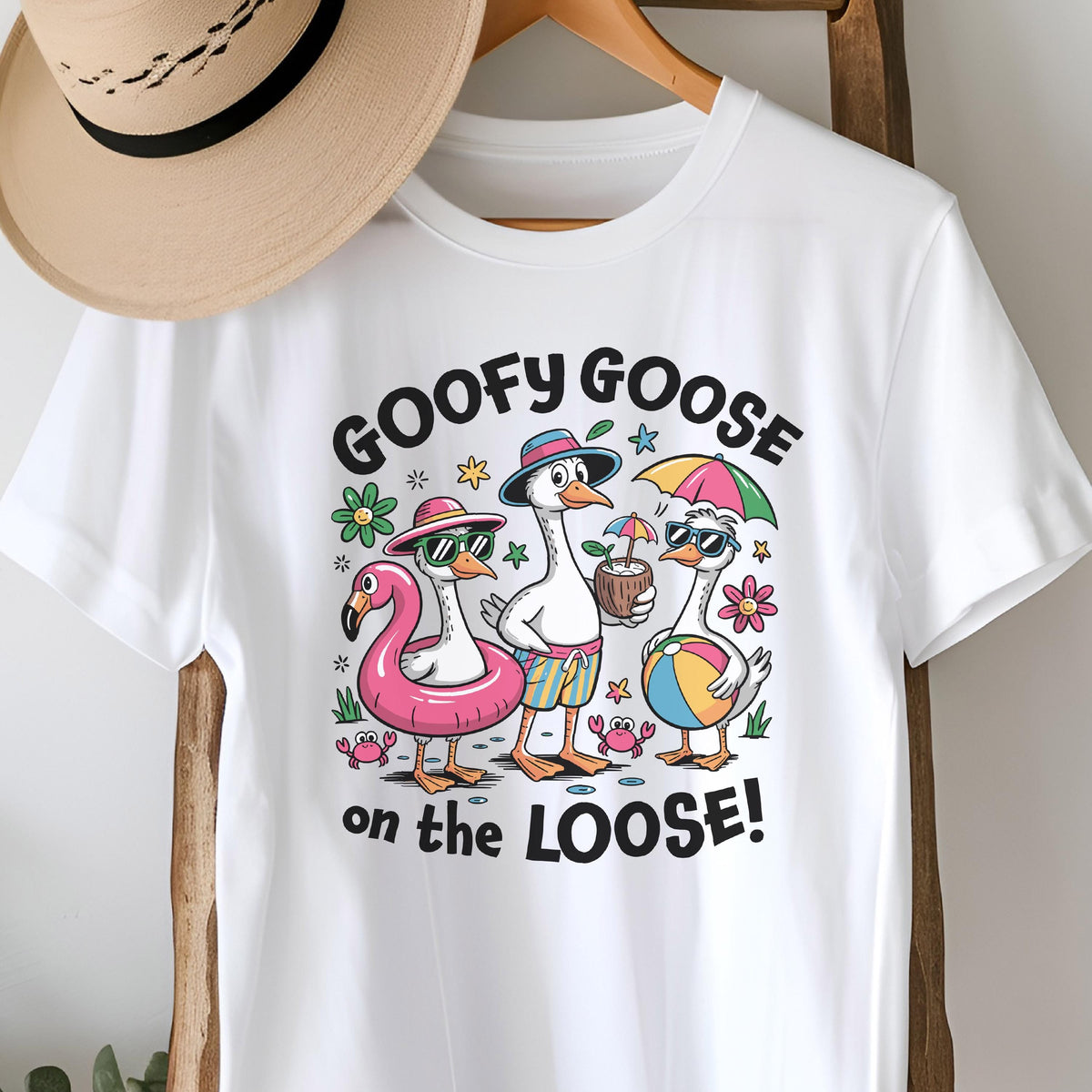 May include: White t-shirt featuring a cartoon design of three geese enjoying a beach day. The geese are wearing sunglasses, hats, and holding beach items. The text "GOOFY GOOSE on the LOOSE!" is printed above and below the geese.