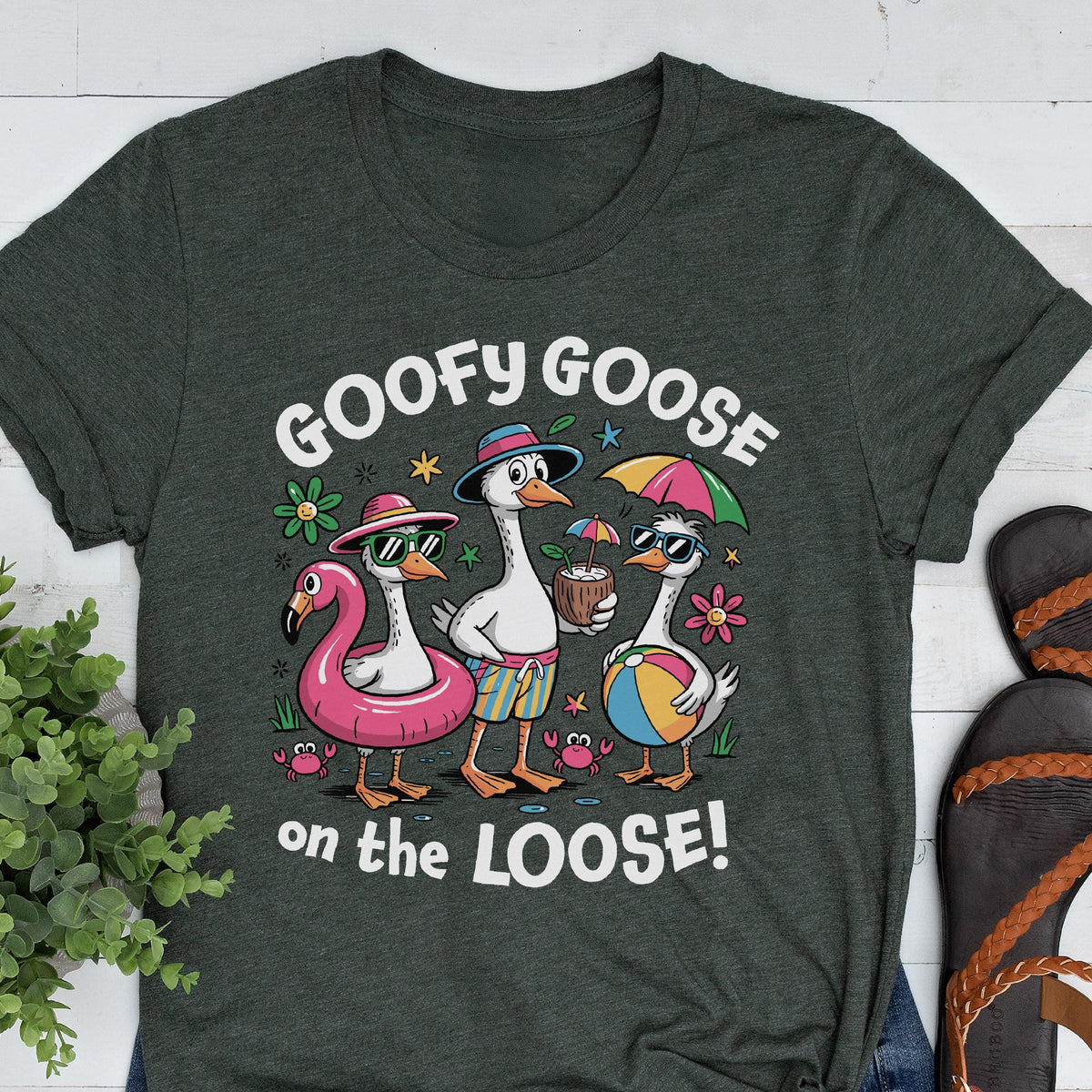 May include: Dark gray t-shirt featuring a graphic of three cartoon geese at the beach. The geese wear sunglasses, hats, and swim trunks, with a flamingo floatie, a coconut drink, and a beach ball. Text reads "GOOFY GOOSE on the LOOSE!"