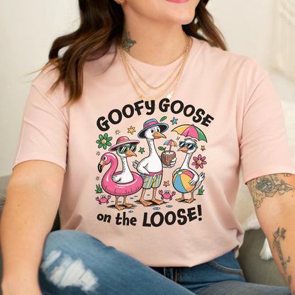 May include: A light pink t-shirt with the text "GOOFY GOOSE on the LOOSE!" features a cartoon illustration of three geese at the beach. One goose wears sunglasses and a flamingo floatie, another holds a coconut drink, and the third holds a beach ball.