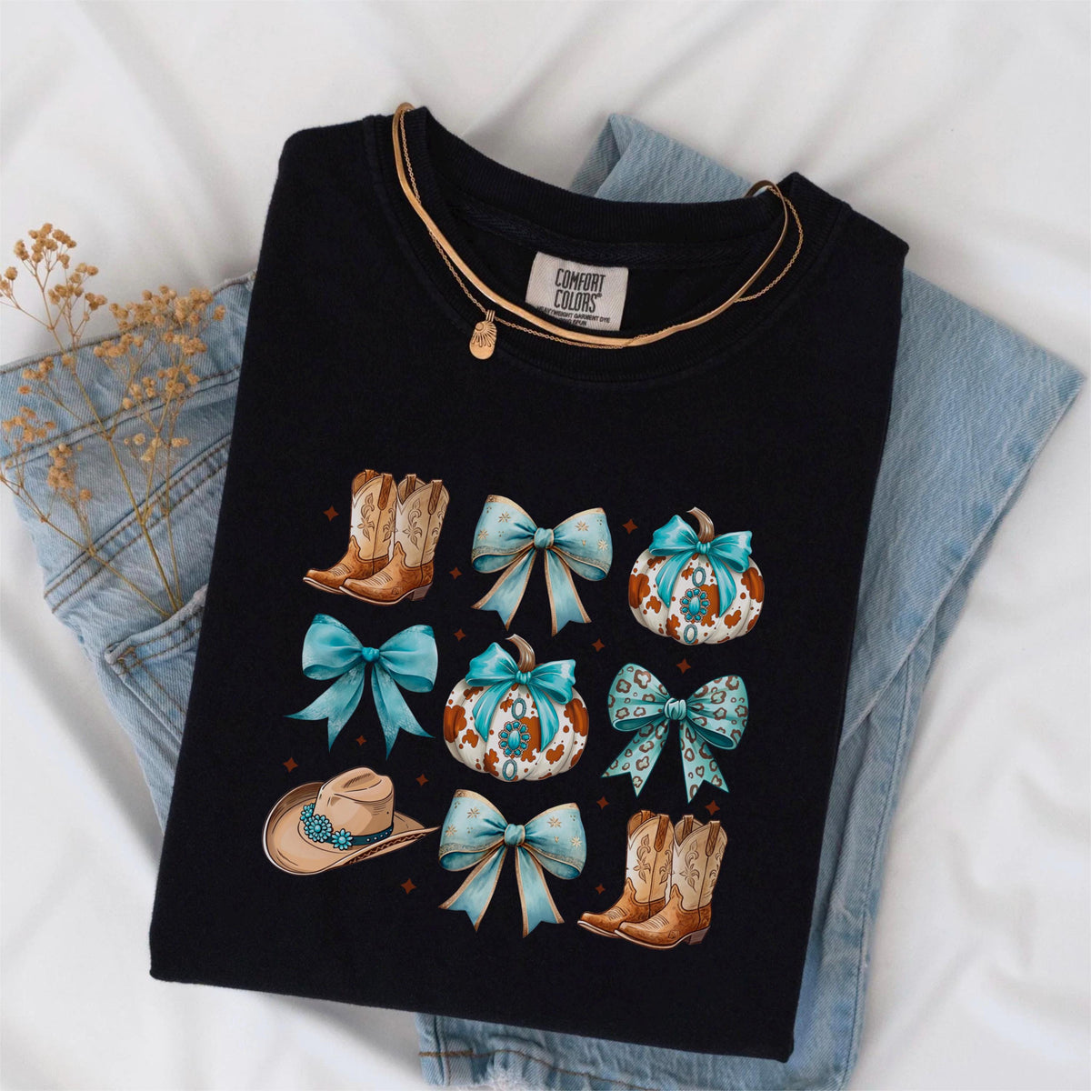May include: Black Comfort Colors t-shirt featuring a design with cowboy boots, turquoise bows, pumpkins, and a cowboy hat. A gold necklace is draped across the shirt. The shirt is laid on top of a pair of blue jeans.