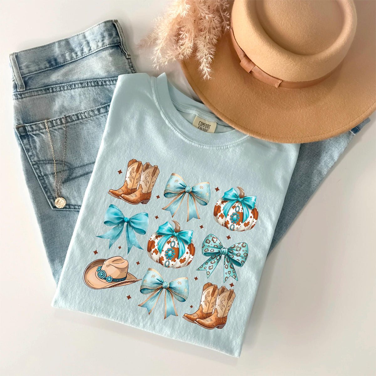 May include: A light blue t-shirt featuring a design of cowboy boots, bows, and pumpkins. The shirt is paired with light wash denim jeans, a tan hat, and a necklace. The overall theme is country and western.