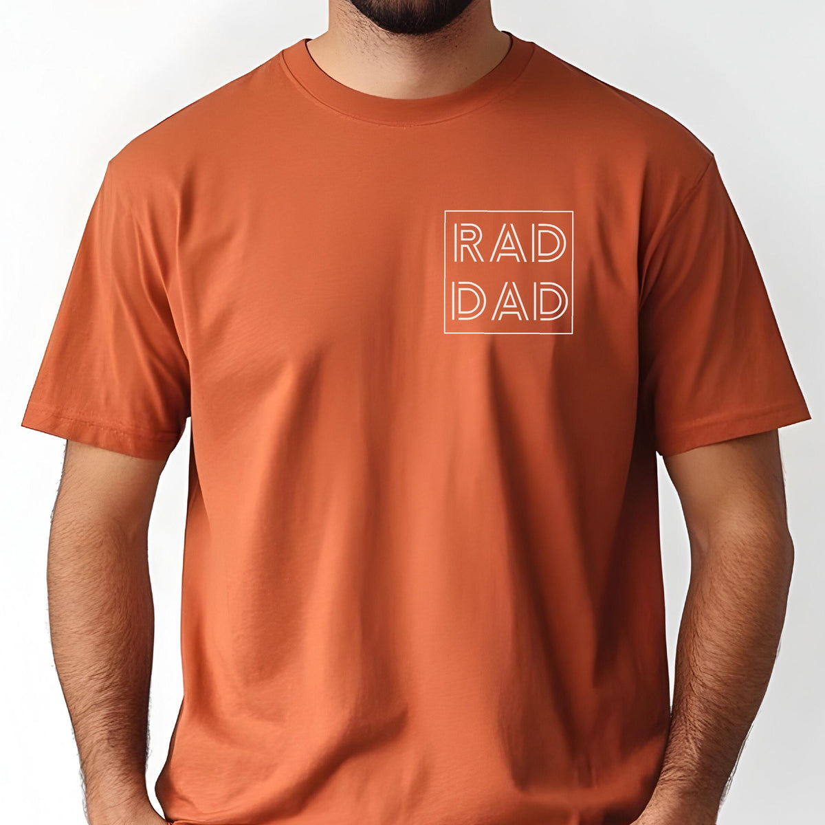 May include: A rust-colored t-shirt with short sleeves. The shirt features the text "RAD DAD" in a white, outlined font, enclosed within a square on the upper left chest area. The shirt is made of a soft material.
