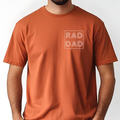 May include: A rust-colored t-shirt with short sleeves. The shirt features the text "RAD DAD" in a white, outlined font, enclosed within a square on the upper left chest area. The shirt is made of a soft material.