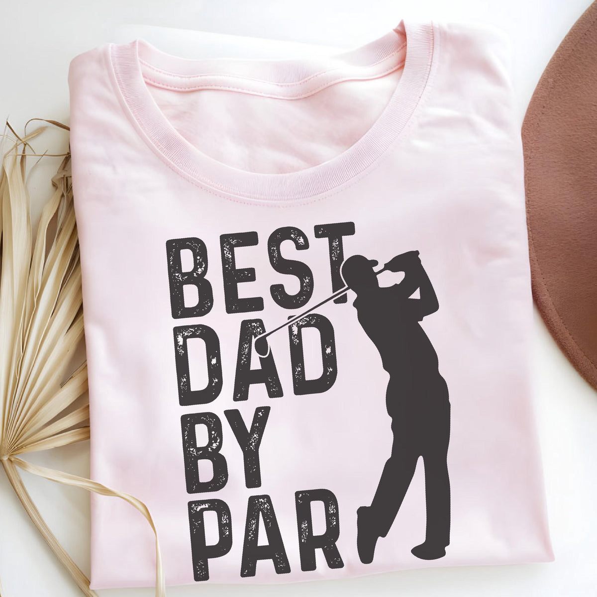 May include: A light pink t-shirt featuring a black graphic print of a golfer in a swing pose. The text "BEST DAD BY PAR" is printed in a bold, distressed black font to the left of the golfer silhouette. The shirt has a crew neckline.