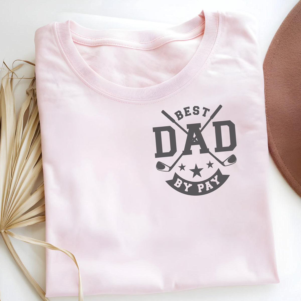 May include: A light pink t-shirt with the text "BEST DAD BY PAY" in dark gray. The design includes crossed golf clubs and stars. A brown hat is partially visible in the background.