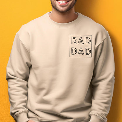 May include: A beige crew neck sweatshirt with the words "RAD DAD" in a black, minimalist font inside a black square. The sweatshirt is made of a soft material.