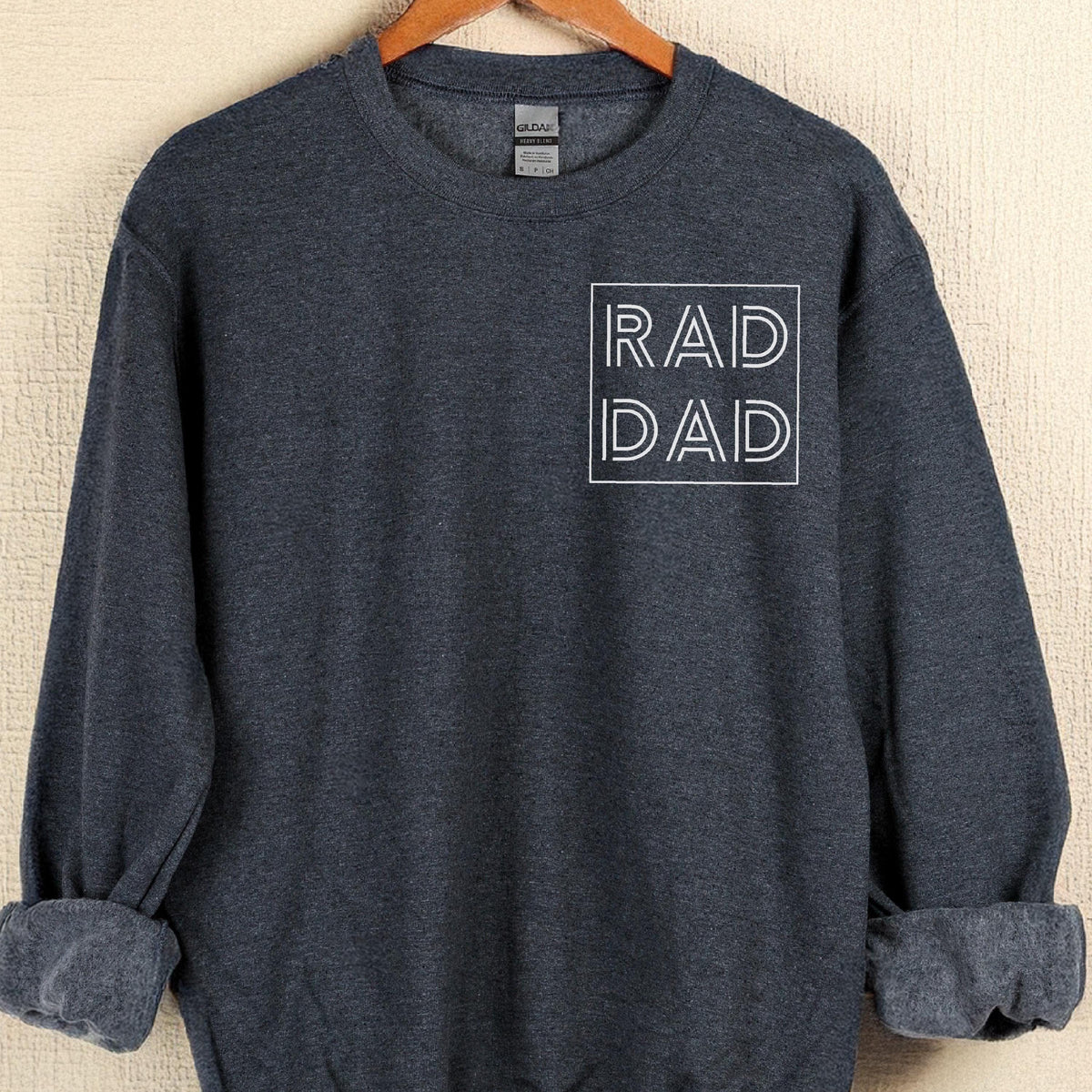 May include: A dark gray sweatshirt with the words "RAD DAD" in a white, outlined font. The sweatshirt is hanging on a wooden hanger. The sleeves are cuffed.