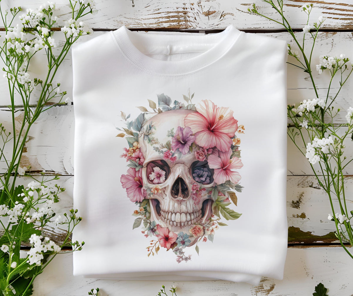 May include: White crewneck sweatshirt featuring a watercolor skull design adorned with pink, purple, and blue flowers. The floral skull graphic is centered on the front of the folded garment, creating a unique and artistic aesthetic.