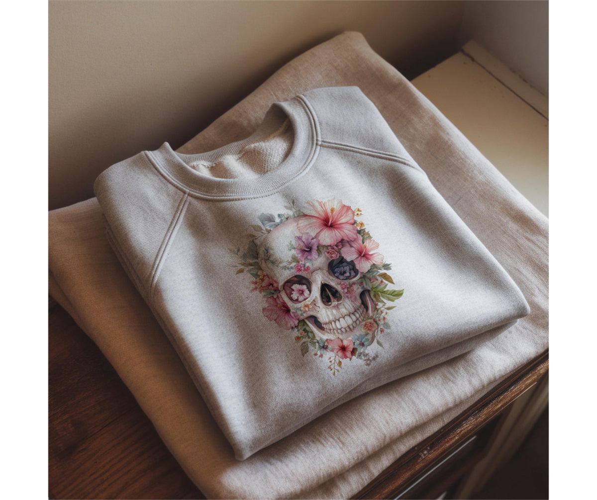 May include: A light gray sweatshirt featuring a floral skull design. The skull is adorned with pink, purple, and blue flowers and green leaves. The sweatshirt is folded on a light brown surface.