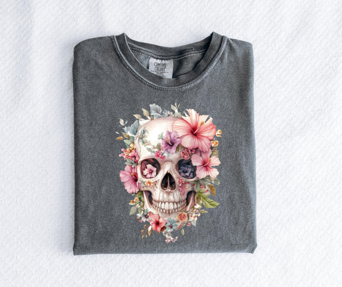 May include: A gray t-shirt featuring a watercolor design of a skull adorned with pink, purple, and white flowers. The skull's eye sockets are filled with floral details, and the design is centered on the shirt.