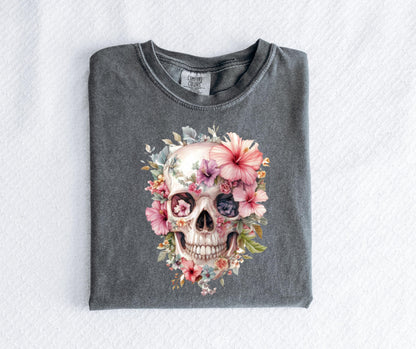 May include: A gray t-shirt featuring a watercolor design of a skull adorned with pink, purple, and white flowers. The skull's eye sockets are filled with floral details, and the design is centered on the shirt.