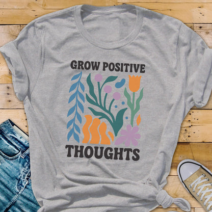 May include: A light gray t-shirt with the words "GROW POSITIVE THOUGHTS" in black. The shirt features a colorful floral design with blue, orange, green, and purple elements. The shirt is made of a soft material.