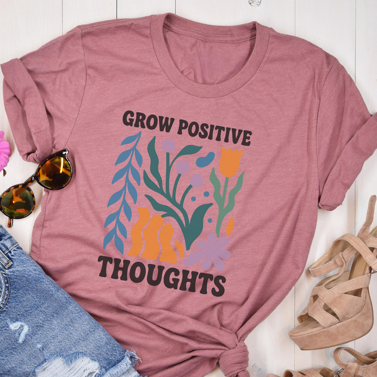 May include: Dusty rose-colored t-shirt with the words "GROW POSITIVE THOUGHTS" in black. The shirt features a floral design in blue, orange, and purple. Sunglasses and denim shorts are next to the shirt.
